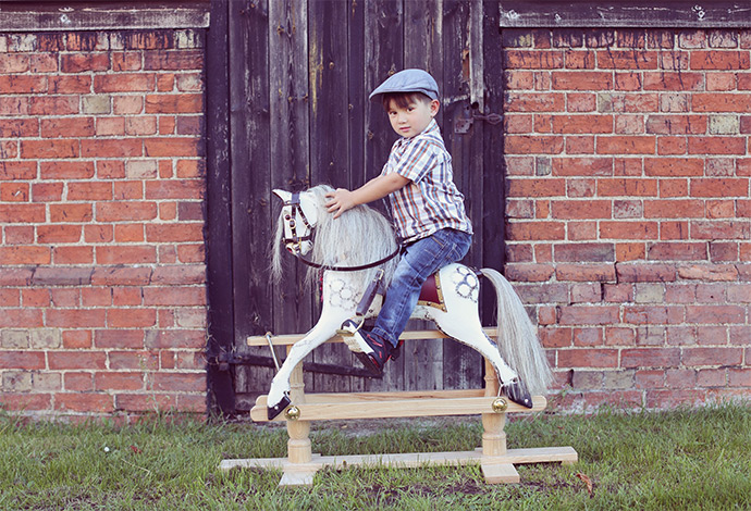 A small boy riding a small dapple grey rocking horse outside